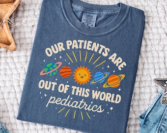 May include: A blue Comfort Colors t-shirt with the text "OUR PATIENTS ARE OUT OF THIS WORLD pediatrics" and a graphic of planets and a smiling sun. The shirt is folded on a white surface.