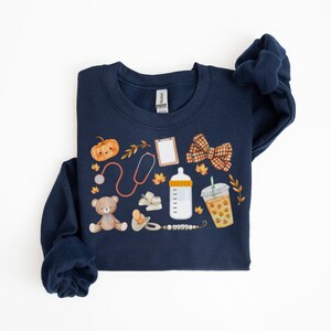May include: A navy blue sweatshirt with a variety of fall-themed baby items printed on it, including a pumpkin, a teddy bear, a baby bottle, a pacifier, a stethoscope, a bow, a coffee cup, and a clipboard.