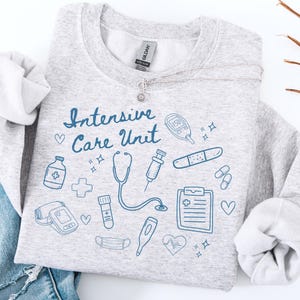 May include: A light gray sweatshirt with the words "Intensive Care Unit" in blue script, along with medical-themed illustrations. A silver necklace with a flower pendant is draped across the sweatshirt.
