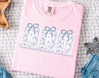 May include: A pale pink t-shirt featuring the letters "RBT" in white, framed by floral patterns and grey bows. The words "Registered Behavior Technician" are printed below. The shirt has a Comfort Colors label.