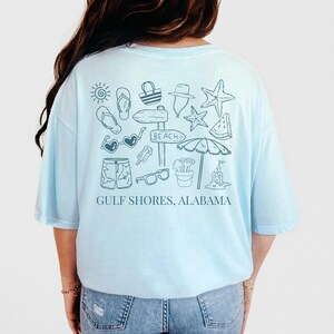 May include: Light blue t-shirt featuring a beach-themed graphic in light blue. The design includes a sun, flip-flops, sunglasses, shorts, and a sign that says "Beach". The text "Gulf Shores, Alabama" is printed below the graphic.