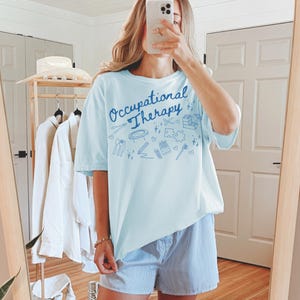 May include: A light blue t-shirt with the text "Occupational Therapy" in blue lettering. The shirt has a variety of blue illustrations of occupational therapy tools and activities.