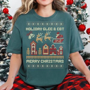 May include: Teal green t-shirt with a festive Christmas design. The shirt features the text "HOLIDAY GLEE & CBT" above a graphic of Santa in a sleigh pulled by reindeer, and the words "MERRY CHRISTMAS" below.