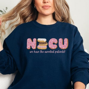 Pode incluir: Uma camisola azul-marinho com o texto "NICU we have the sweetest patients!" em rosa. As letras s&atilde;o compostas por donuts cor-de-rosa com granulado rosa e branco.