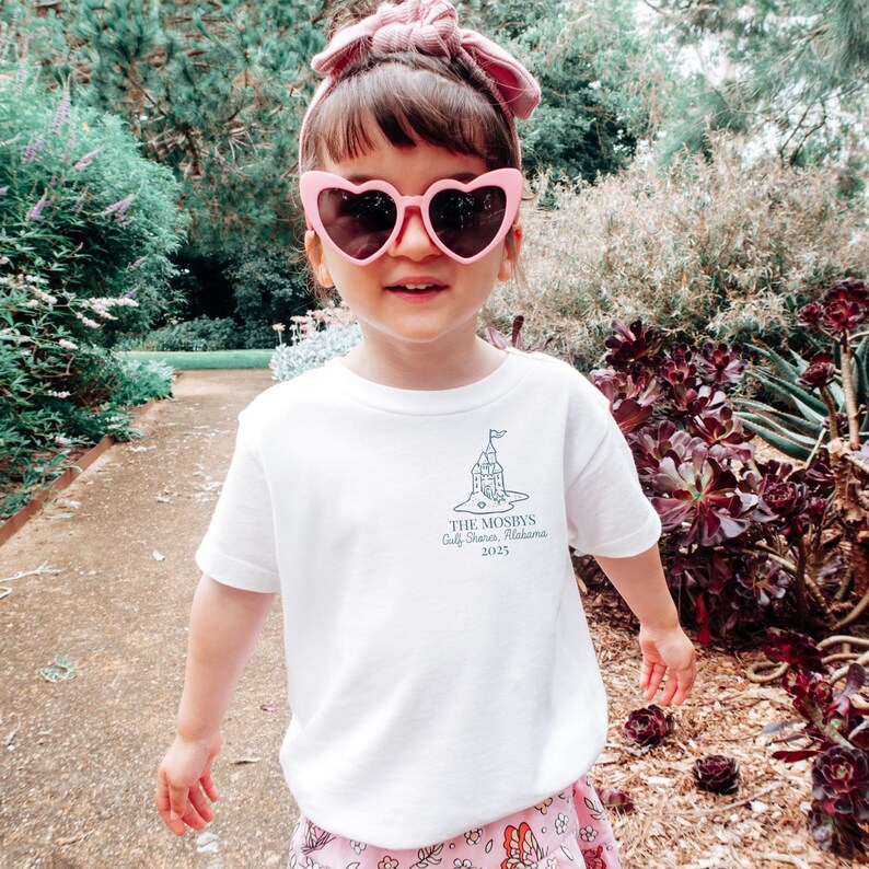 May include: A young child wearing a white t-shirt with a castle graphic and text, pink heart-shaped sunglasses, and a pink headband. The text on the shirt reads "THE MOSBYS Gulf Shores, Alabama 2025". The child is smiling outdoors.