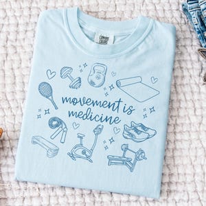 May include: Light blue t-shirt with a graphic design of various fitness equipment and the text "movement is medicine".