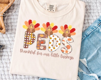 Può includere: T-shirt color crema con grafica a tema Ringraziamento. La parola "PEDS" è visualizzata con illustrazioni di tacchini sopra ogni lettera. Il testo "thankful for our little turkeys" è sotto.