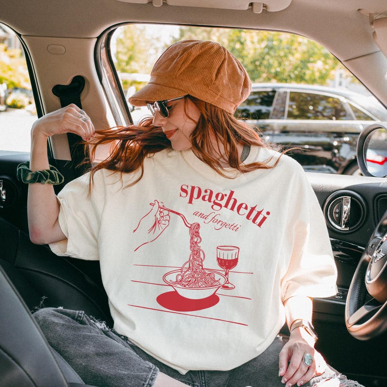 May include: A white t-shirt with a red graphic of a hand holding a fork and a plate of spaghetti. The text "spaghetti and forgetti" is written in red above the graphic.