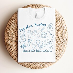 May include: White t-shirt with a blue design that says "Pediatric Oncology" and includes illustrations of medical supplies, a teddy bear, and balloons. The text below the design says "play is the best medicine."