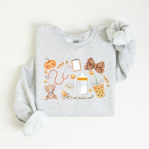 May include: A gray sweatshirt with a fall-themed design featuring a pumpkin, stethoscope, baby bottle, teddy bear, pacifier, bow, and a cup of coffee. The design is in watercolor style with orange, brown, and yellow colors.