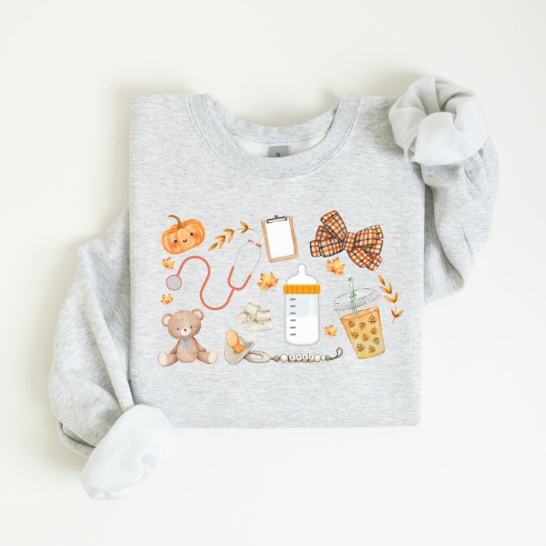 May include: A gray sweatshirt with a fall-themed design featuring a pumpkin, stethoscope, baby bottle, teddy bear, pacifier, bow, and a cup of coffee. The design is in watercolor style with orange, brown, and yellow colors.