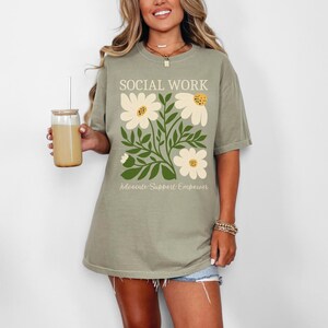 May include: A sage green t-shirt with a floral design and the text "SOCIAL WORK Advocate. Support. Empower."