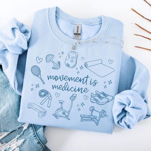 Pode incluir: Camisola azul claro com o texto "movement is medicine" em letras azuis. A camisola apresenta uma variedade de &iacute;cones de fitness, incluindo um kettlebell, halteres, um tapete de ioga, uma corda de saltar, uma bicicleta de exerc&iacute;cio, um banco de muscula&ccedil;&atilde;o, sapatos de corrida e uma raquete de t&eacute;nis.