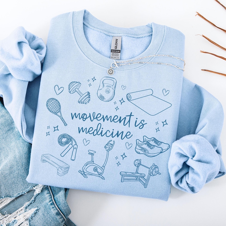 Pode incluir: Camisola azul claro com o texto "movement is medicine" em letras azuis. A camisola apresenta uma variedade de &iacute;cones de fitness, incluindo um kettlebell, halteres, um tapete de ioga, uma corda de saltar, uma bicicleta de exerc&iacute;cio, um banco de muscula&ccedil;&atilde;o, sapatos de corrida e uma raquete de t&eacute;nis.