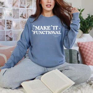 May include: A dusty blue long-sleeve crewneck sweatshirt featuring the white text "MAKE IT FUNCTIONAL" in a retro-style font, accompanied by small star accents and the words "OCCUPATIONAL THERAPY" printed in a smaller font underneath.