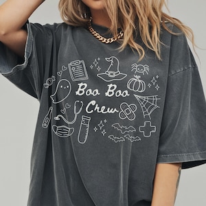 May include: Dark gray t-shirt with the phrase "Boo Boo Crew" and Halloween-themed illustrations in white. The design includes a ghost, witch hat, spiderweb, pumpkin, and medical symbols. The shirt has short sleeves and a relaxed fit.