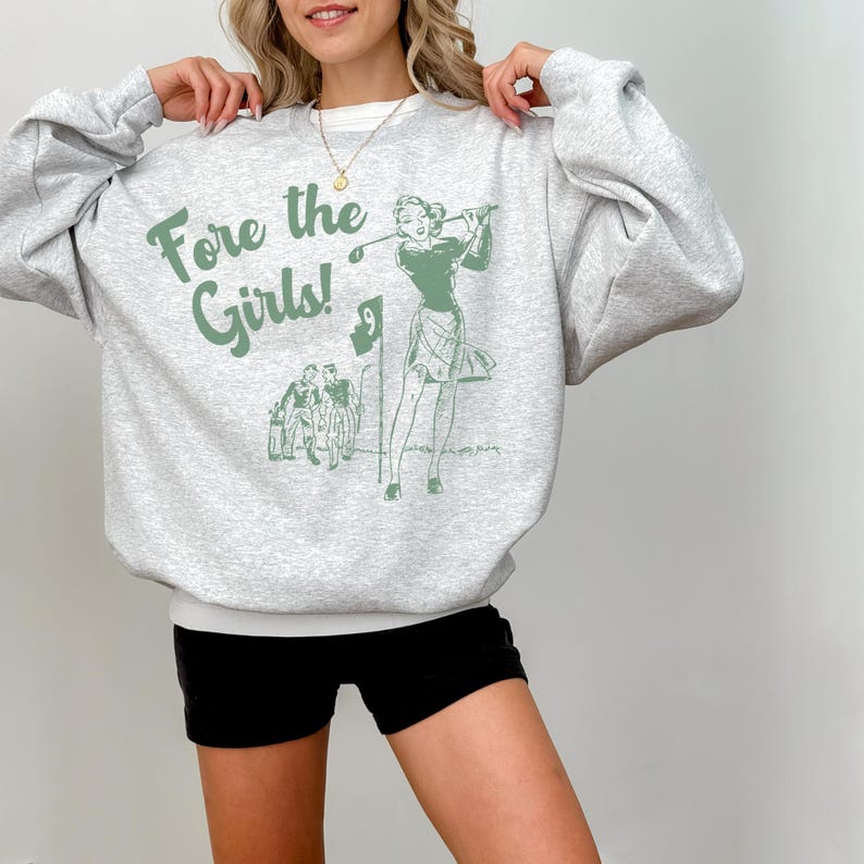 K&ouml;nnte beinhalten: Ein graues Sweatshirt mit dem Text "Fore the Girls!" und einer gr&uuml;nen Illustration einer Frau, die Golf spielt.