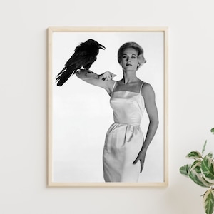 May include: Black and white photograph of a woman in a white dress holding a black raven on her shoulder. The photo is framed and hanging on a white wall.