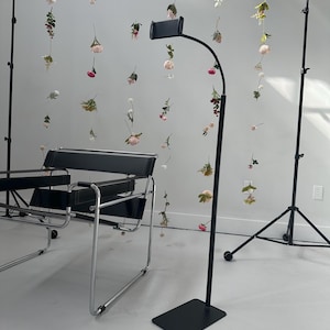 Puede incluir: Un soporte de suelo negro ajustable con una base rectangular y un brazo curvo, que sostiene un dispositivo negro. El fondo presenta arreglos florales colgantes y una moderna silla negra.