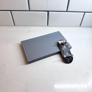 May include: A miniature skateboard with a detailed graphic design rests on a gray, rectangular platform with a brick-like border. The skateboard features a black deck with a colorful design and small wheels.