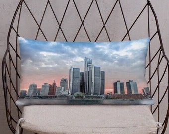 May include: A decorative pillow featuring a panoramic view of the Detroit skyline at sunset. The image shows a cluster of skyscrapers against a cloudy sky with a pink and orange hue.