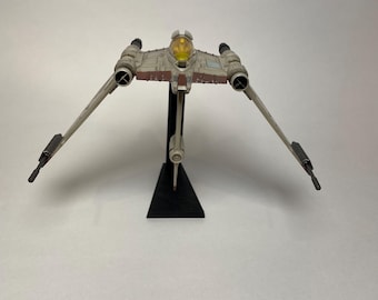 May include: A white and brown Star Wars B-Wing fighter spaceship model with a yellow cockpit canopy. The model is mounted on a black stand.