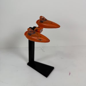 May include: An orange toy spacecraft with a black stand. The spacecraft has a sleek, aerodynamic design with a cockpit and wings. The stand is rectangular and supports the spacecraft, which is angled upwards.