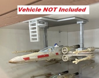 May include: A white plastic model of a Star Wars X-Wing fighter suspended from a ceiling mount. The mount is made of gray plastic and has a ladder attached to it. The text "Vehicle NOT Included" is visible in the image.