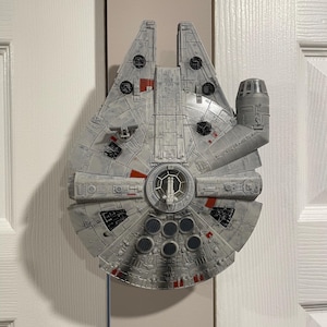May include: A detailed model of the Millennium Falcon starship, featuring a gray and white color scheme with red and black accents. The model has intricate detailing, including various panels, turrets, and a central cockpit. It is mounted on a white door.