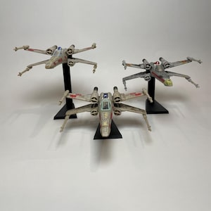 May include: Three Star Wars X-Wing fighter model kits, each painted in a different color scheme. The first is a light gray with red and white markings, the second is a dark gray with red and white markings, and the third is a light gray with red and white markings. All three models are on black stands.