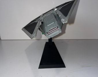 May include: A gray and black model of a TIE fighter spacecraft, a fictional starship from the Star Wars franchise. The model is mounted on a black stand, with the wings angled upward. The spacecraft has detailed paneling and a cockpit.