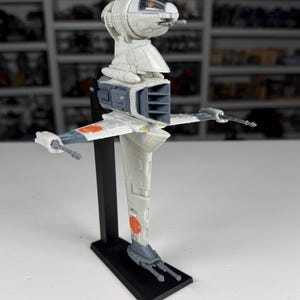 May include: A detailed model of a Star Wars B-wing starfighter, predominantly white with gray and orange accents. The model is mounted on a black stand, showcasing its distinctive wing design and weaponry. A collectible item.