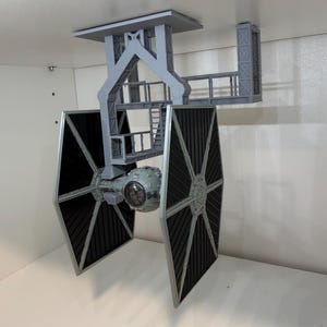 May include: A gray, 3D-printed model of a TIE fighter from Star Wars, mounted to a ceiling fixture. The fighter has black solar panels and a detailed cockpit. The mounting structure is also gray and features a platform and support beams.