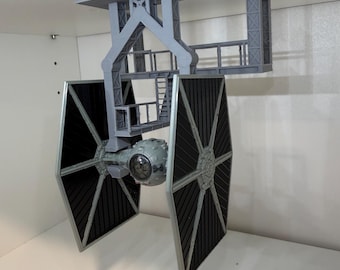 May include: A gray, 3D-printed model of a TIE fighter from Star Wars, mounted to a ceiling fixture. The fighter has black solar panels and a detailed cockpit. The mounting structure is also gray and features a platform and support beams.