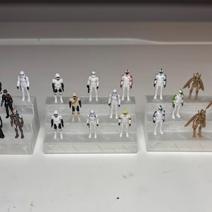 May include: A collection of miniature Star Wars figurines, including Stormtroopers, Darth Vader, and other characters. The figures are arranged on clear, tiered display stands, showcasing a variety of colors and designs. The scene is set against a plain white background.