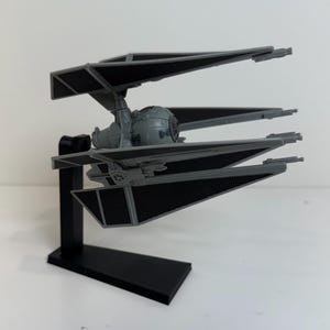 May include: A gray model of a TIE fighter spacecraft, a fictional starship from the Star Wars franchise. The model has two large solar panels and is mounted on a black stand. The spacecraft is gray with black details.