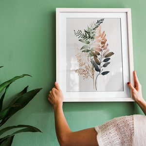 May include: A framed botanical print with watercolour illustrations of leaves in green, brown, and beige tones. The artwork is in a white frame against a green wall, with a plant in the foreground.