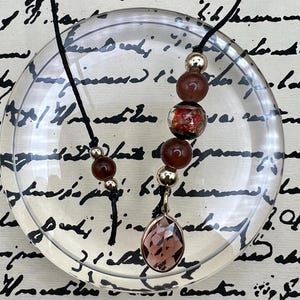 May include: A brown and gold beaded necklace with a large, faceted, purple gemstone pendant. The necklace is on a black cord and is displayed on a clear surface.