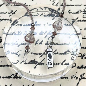 May include: A beaded eyeglass chain featuring small mauve seed beads, clear and mauve heart-shaped beads, and silver-toned accents. One end terminates in a silver-toned rectangular charm with the word LOVE engraved in black lettering.