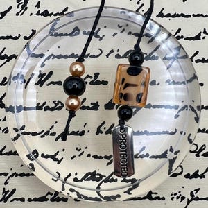 May include: Two beaded pendants on black cords. One features gold and black beads, the other a rectangular amber-colored bead with black spots, a black bead, and a silver tag that reads "PROTECTED".