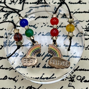 May include: Four friendship charm necklaces. Each necklace has a black cord, colorful beads, and a charm with a rainbow and the words "Best Friends". The charms are gold-colored.