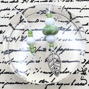 May include: Two white strings featuring green and white beads. One string has a silver-toned metal leaf charm attached at the bottom. The beads are a mix of small green cylindrical beads and larger smooth white spherical beads.