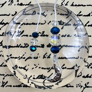 May include: Two silver crescent moon charms on white strings with blue and clear glass beads. The charms are hanging from a clear glass plate.