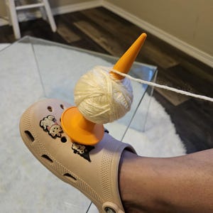 May include: A light beige clog shoe featuring two small cartoon character charms on the top. An orange plastic yarn spindle is inserted into the shoe, holding a ball of cream-colored yarn that is being pulled from the side.
