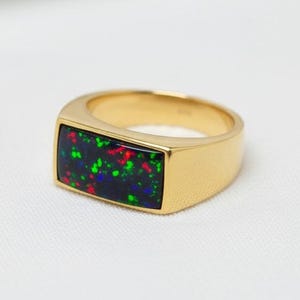 May include: A gold ring with a rectangular black opal stone. The opal features a vibrant display of green, red, and blue colors. The ring has a polished finish and a classic, elegant design.