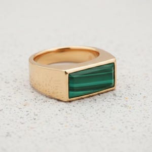 Può includere: Un anello color oro con una pietra rettangolare di malachite verde. L'anello ha una fascia spessa e una superficie strutturata. La pietra di malachite è di un verde vibrante con strisce verde scuro.