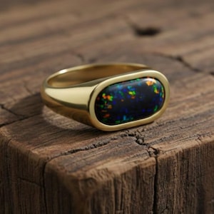 May include: A gold ring featuring a rectangular black opal gemstone. The opal displays flashes of blue, green, and orange. The ring is set on a dark wooden surface.