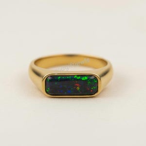 Puede incluir: Un anillo de oro con una piedra preciosa de ópalo negro rectangular. El ópalo muestra un vibrante juego de colores con toques de verde, rojo y azul. El anillo tiene un acabado liso y pulido.