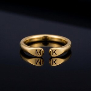 Peut inclure: Une bague ouverte de couleur or avec les lettres "M" et "K" gravées sur le devant. La bague est posée sur un fond sombre, et les lettres sont clairement visibles. Il s'agit d'un bijou.