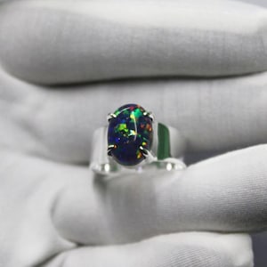Pode incluir: Anel de prata com uma opala oval. A opala apresenta um jogo de cores vibrante, com flashes de verde, azul e amarelo sobre um fundo escuro. O anel tem uma banda de prata polida e simples.